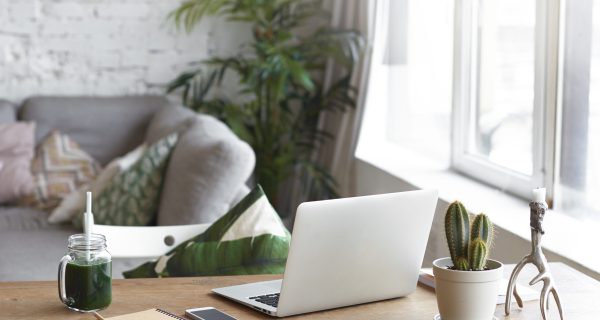 Workplace of modern freelancer leading healthy lifestyle: open generic laptop computer, cactus in pot, smart phone, copybook and glass of green smoothie on wooden desk in stylish home office interior